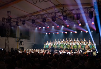 Szeroki widok na podświetloną scenę na której znajdują się Artyści Chóru w kostiumach łowickich, Artyści Orkiestry w kostiumach wilanowskich wraz z Dyrygentem