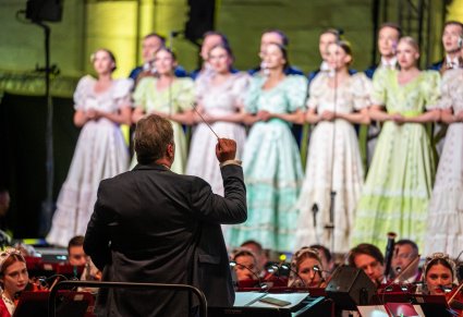 Na pierwszym planie Dyrektor Jacek Boniecki zwrócony w stronę Orkiestry i Chóru. Chór w kostiumach stylowych z końca XIX w.
