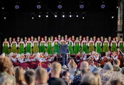 Widok przez publiczność na scenę plenerową. Na pierwszym planie Dyrygent w garniturze zwrócony w stronę Artystów Chóru w kostiumach kurpiowskich, niżej sceny Artyści Orkiestry w kostiumach wilanowskich podczas grania na instrumentach