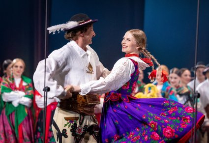 Na pierwszym planie para Artystów w kostiumach podhalańskich podczas wykonywania choreografii tanecznej w stylu góralskim
