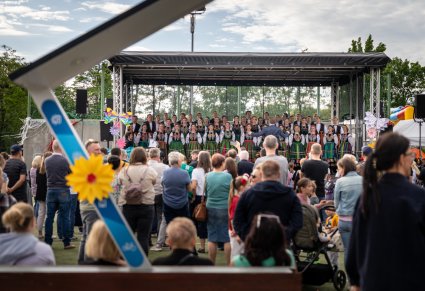 Szeroki widok na publiczność i scenę. Na scenie Dyrygent zwrócony w stronę Artystów Chóru w kostiumach łowickich, widoczny również Akompaniator grający na instrumencie