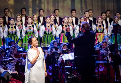 Na pierwszym planie Solistka w długiej, białej sukni obok zwrócony do Orkiestry Dyrektor Jacek Boniecki, który dyryguje koncert w tle chór w kostiumach łowickich