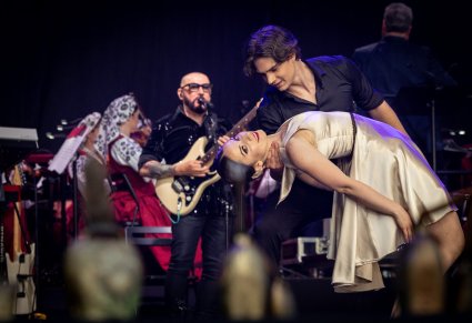Na pierwszym planie para Artystów Baletu w kostiumach współczesnych podczas wykonywania choreografii tanecznej. Na drugim planie Solista zespołu Kombii podczas śpiewu, za nim Orkiestra zespołu "Mazowsze" w kostiumach wilanowskich
