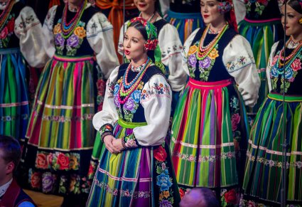 Zbliżenie na Solistkę w trakcie śpiewu, w tle część Artystów Chóru w kostiumach łowickich