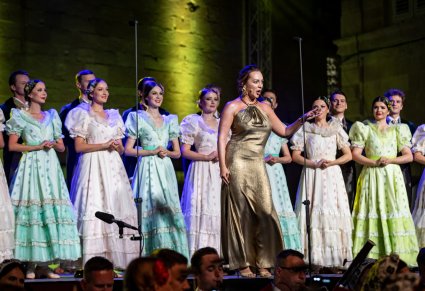 Zbliżenie na śpiewającą solistkę w długiej, złotej sukni a za nią Artyści z chóru w kostiumach stylowych z końca XIX W.