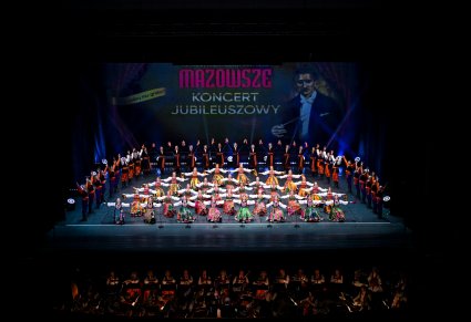 Na scenie Artyści w kostiumach ludowych podczas finałowej choreografii tanecznej, niżej część orkiestry w kostiumach wilanowskich