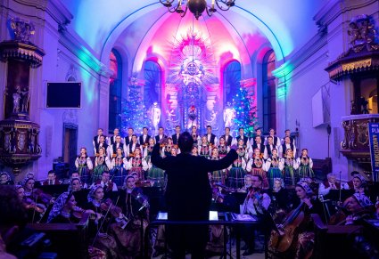 Na pierwszym planie maestro a w tle orkiestra oraz chór. Kolorowe światła oświetlają wnętrze kościoła nadając scenie niepowtarzalny klimat