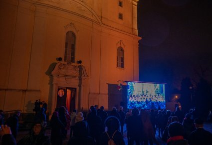 Wejście do kościoła w Pile. Przed kościołem stoi wielki ekran, na którym wyświetlany jest koncert kolęd w wykonaniu zespołu "Mazowsze". Koncert ogląda zgromadzony tłum.