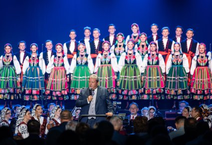 Chór w kostiumach łowickich stoi na scenie a przed nimi orkiestra i przemawiający dyrygent