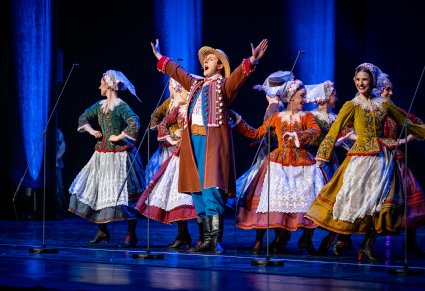 Na pierwszym planie śpiewający Artysta w kostiumie rzeszowskim a za nim tancerki wykonujące choreografię taneczną