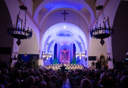 Widoczna cała przestrzeń kościelna, większość publiki, ołtarz przed którym stoją Artyści Chóru w kostiumach łowickich a przed nimi Orkiestra wraz z Dyrektorem Jackiem Bonieckim, który dyryguje koncert