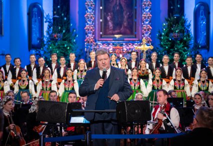 Dyrektor Jacek Boniecki zwrócony w stronę publiczności, przemawiający do mikrofonu. Za nim Orkiestra i Artyści Chóru w kostiumach łowickich