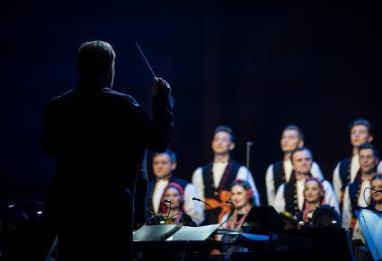 Na pierwszym planie Dyrektor Jacek Boniecki zwrócony w stronę Orkiestry i Artystów Chóru w kostiumach łowickich