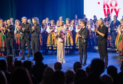 Anna Wyszkoni w złotej sukni trzymajaca bukiet kwiatów na scenie z Artystami "Mazowsza" i ze swoim zespołem