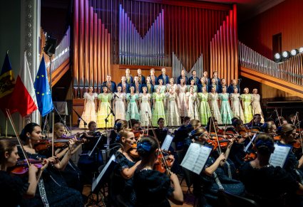 Na pierwszym planie część orkiestry w stylowych kostiumach a za nią na scenie Artyści chóru w stylowych kostiumach z XIX w.