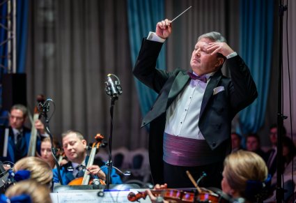 Zbliżenie na Dyrektora Jacka Bonieckiego który w górze trzyma batutę, widać część orkiestry