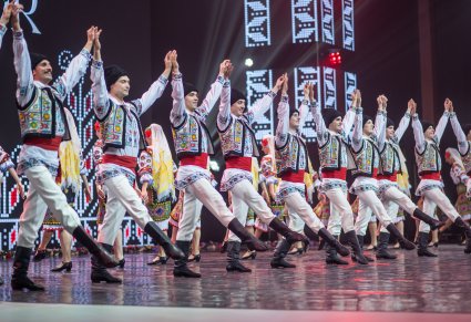 Grupa Artystów z zespołu Baletul Național JOC w kostiumach ludowych w trakcie wykonywania choreografii tanecznej na scenie