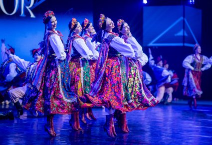 Na pierwszym planie Artystki w kostiumach krakowskich z zespołu "Mazowsze" na scenie w trakcie wykonywania choreografii tanecznej