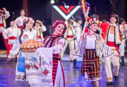 Zbliżenie na Artystki z zespołu Baletul Național JOC w kostiumach ludowych w trakcie wykonywania choreografii tanecznej, Artystka na pierwszym planie trzyma okrągły chleb