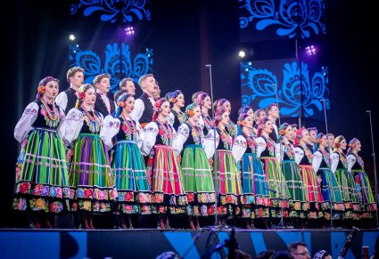 Zbliżenie na chór zespołu "Mazowsze" w kostiumach łowickich w trakcie śpiewu na scenie