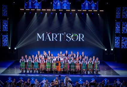 Na scenie Chór zespołu "Mazowsze" wraz z Solistą w kostiumach łowickich w trakcie śpiewu, widoczna część orkiestry