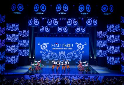 Na scenie Artyści zespołu "Mazowsze" w kostiumach łowickich wykonujący choreografię taneczną, widoczna orkiestra