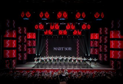 Na scenie Artyści Baletul Național JOC w kostiumach ludowych wykonujący choreografię taneczną, widoczna część orkiestry z obu zespołów