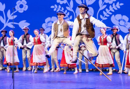 Na pierwszym planie dwóch Artystów z zespołu "Mazowsze" w kostiumach jurgowskich w trakcie wykonywania akrobacji tanecznych, na drugim planie część z zespołu również w kostiumach jurgowskich