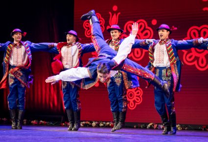 Na pierwszym planie Artysta z zespołu "Mazowsze" w kostiumie podegrodzkim wykonujący spektakularne akrobacje taneczne na scenie