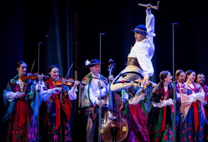 Na pierwszym planie Artysta z zespołu "Mazowsze" w kostiumie podhalańskich wykonujący spektakularną figurę taneczną a za nim na scenie grupa tancerzy i Muzyków również w kostiumach podhalańskich