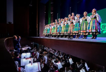 Na scenie Chór zespołu "Mazowsze" w kostiumach łowickich w trakcie śpiewu, widoczna również orkiestra w kostiumach wilanowskich