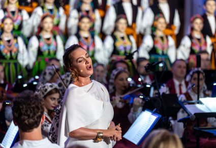 Zbliżenie na Solistkę w białej suknie trakcie śpiewu, za nią Artyści z chóru w kostiumach łowickich