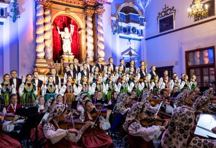 Na pierwszym planie orkiestra a za nią Artyści z chóru w kostiumach łowickich ustawieni w rzędzie. Tłem jest piękny ołtarz
