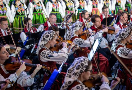 Zbliżenie na część orkiestry smyczkowej, w tle Artyści z chóru w kostiumach łowickich