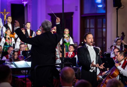 Na pierwszym planie Dyrektor Jacek Boniecki zwrócony w stronę orkiestry, energicznie dyryguje koncert, na drugim planie Solista w trakcie śpiewu ubrany w smoking, za Orkiestrą część chóru w kostiumach łowickich