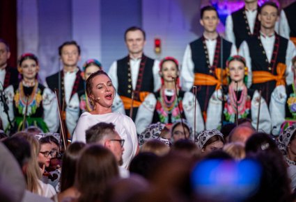 Na pierwszym planie śpiewająca solistka w białej sukience a za nią Artyści chóru w kostiumach łowicki ustawieni w rzędy