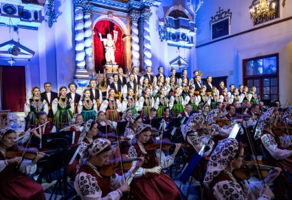 Na pierwszym planie Orkiestra a za nią chór w kostiumach łowickich ustawiony w rzędy tłem "sceny" jest pięknie bogato zdobiony ołtarz