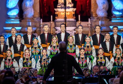 Na pierwszym planie Dyrektor Jacek Boniecki zwrócony w stronę Orkiestry i chóru, który jest w kostiumach łowickich