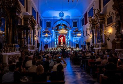 Pokazana przestrzeń świątyni w której odbywa się koncert, na zdjęciu widoczna publiczność a na końcu pomieszczenia przed ołtarzem Orkiestra wraz z chórem