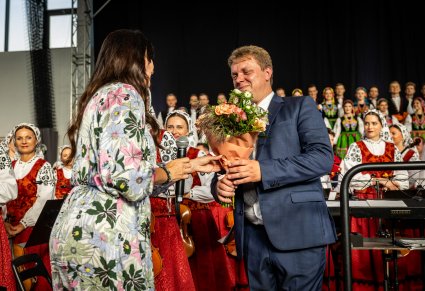 Dyrygent odbierający kwiaty po koncercie, w tle widoczna orkiestra z chórem