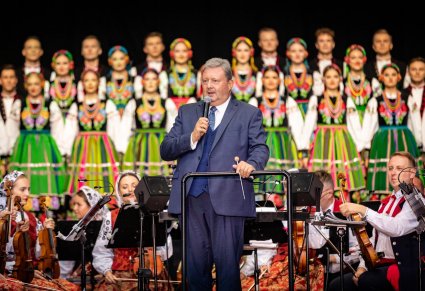 Dyrektor przemawiający przed koncertem, chór i orkiestra widoczne w tle