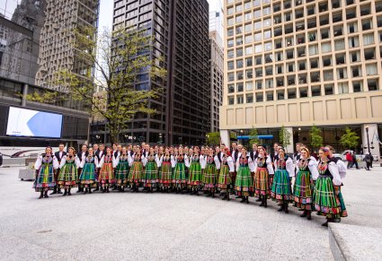 Artyści Chóru w kostiumach łowickich ustawieni w dwóch rzędach w trakcie śpiewania Hymnu Polski w trakcie Ceremonii Podniesienia Podniesienia Flagi