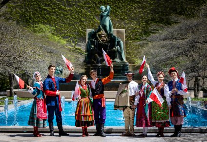 Cztery pary Artystów w kostiumach kujawskich, sannickich, podhalańskich i krakowskich przed fontanną. Artyści trzymają w górze polskie flagi