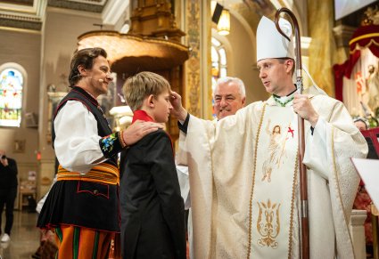Młodzieniec wraz ze swoim świadkiem Artystą zespołu "Mazowsze" w kostiumie łowickim, stoją przed Biskupem, który udziela chłopakowi Bierzmowania