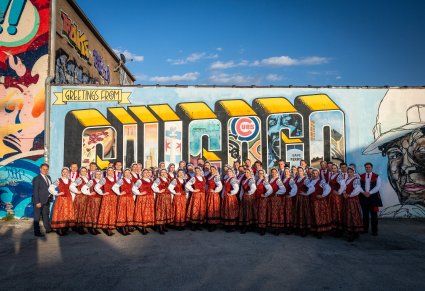 Artyści Orkiestry zespołu "Mazowsze" w kostiumach wilanowskich ustawieni przed muralem z napisem GREETING FROM CHICAGO