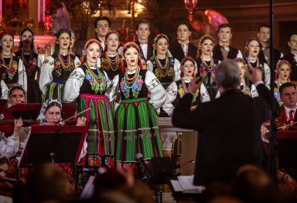 Na pierwszym planie dwie Solistki w tle reszta część Chóru w kostiumach łowickich, widoczna również część Orkiestry w kostiumach wilanowskich wraz z Dyrygentem zwróconym w stronę Chóru i Orkiestry