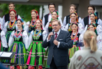 Na pierwszym planie Pan Mirosław Ziomek, wieloletni kierownik chóru zespołu "Mazowsze" podczas przemówienia dla Publiczności, za nim widoczna część Artystów chóru w kostiumach łowickich