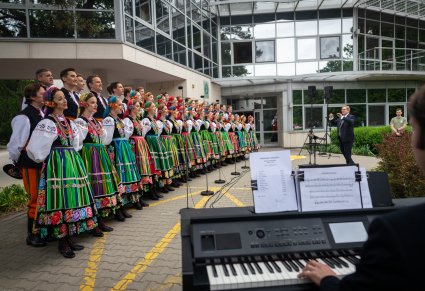 Na pierwszym planie Akompaniator grający na instrumencie, w drugim planie Artyści chóru w kostiumach łowickich wraz z Dyrygentem zespołu