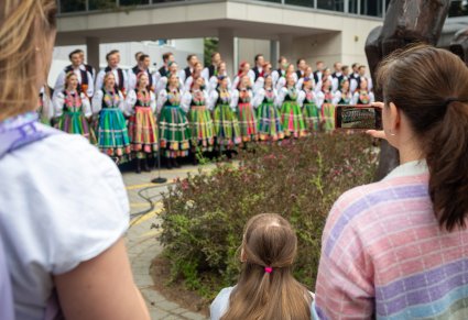 Na pierwszym planie widoczna część Publiczności, która rejestruje telefonem koncert, w tle Artyści chóru w kostiumach łowickich