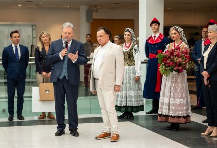 Dyrektor zespołu "Mazowsze" przemawiający do mikrofonu, obok Marszałek Województwa Mazowieckiego a za nimi pracownicy oraz Artyści w kostiumach wilanowskich